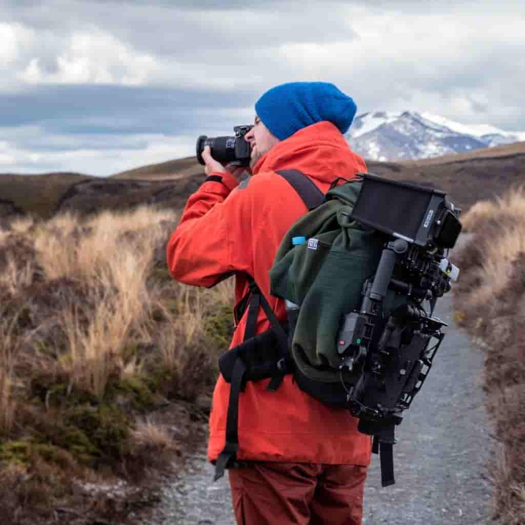 A photographer wearing a camera backpack and taking a photo while standing on a trail near mountains.
