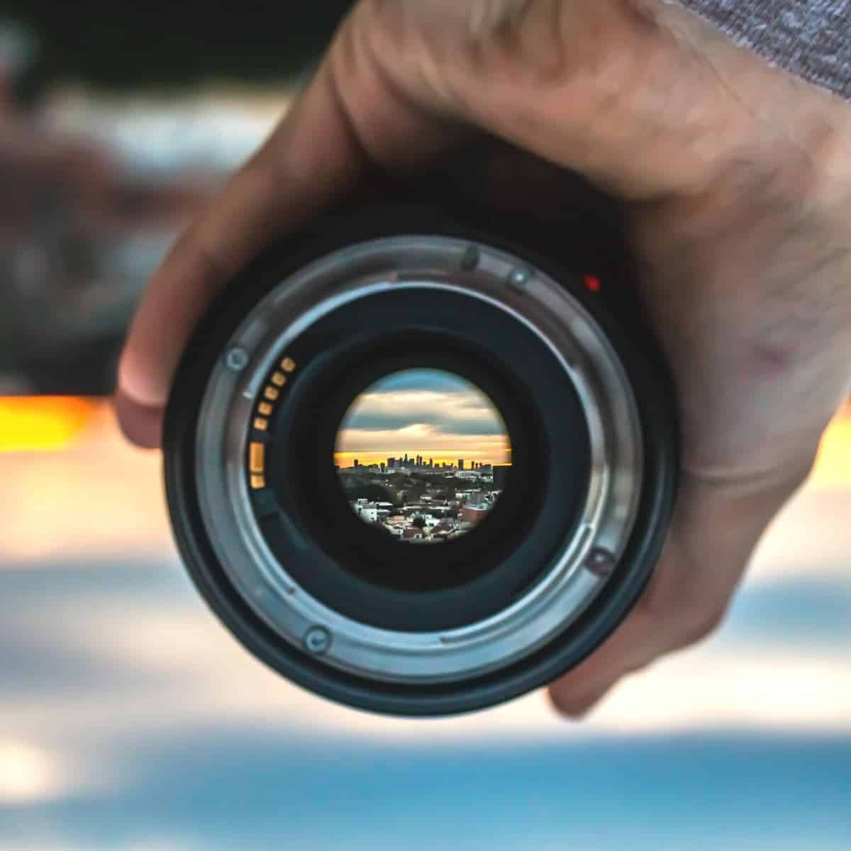 Hand holding a camera lens showing a sunset and city skyline.