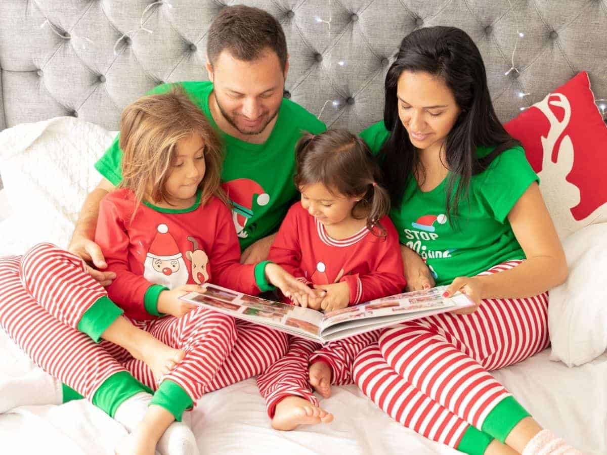 Family wearing matching Christmas pajamas and looking at a book while sitting on a bed.