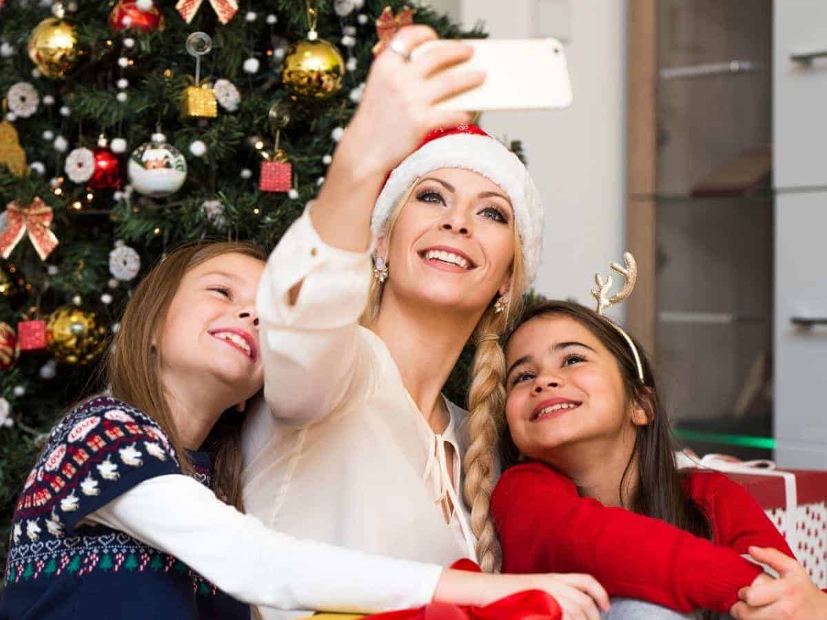 Mom taking selfie with daughters in front of Christmas tree.