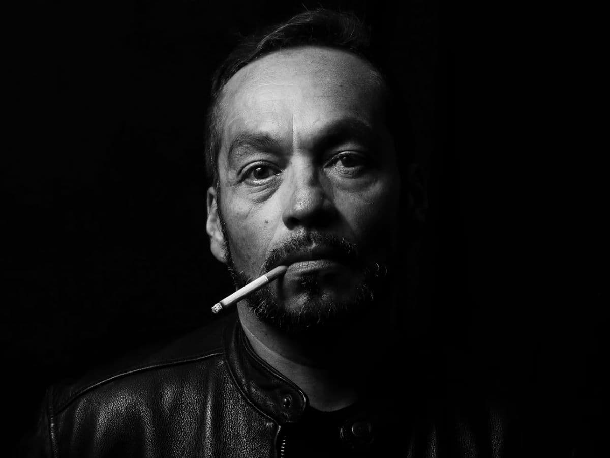 Greyscale headshot of a man with a cigarette in his mouth.