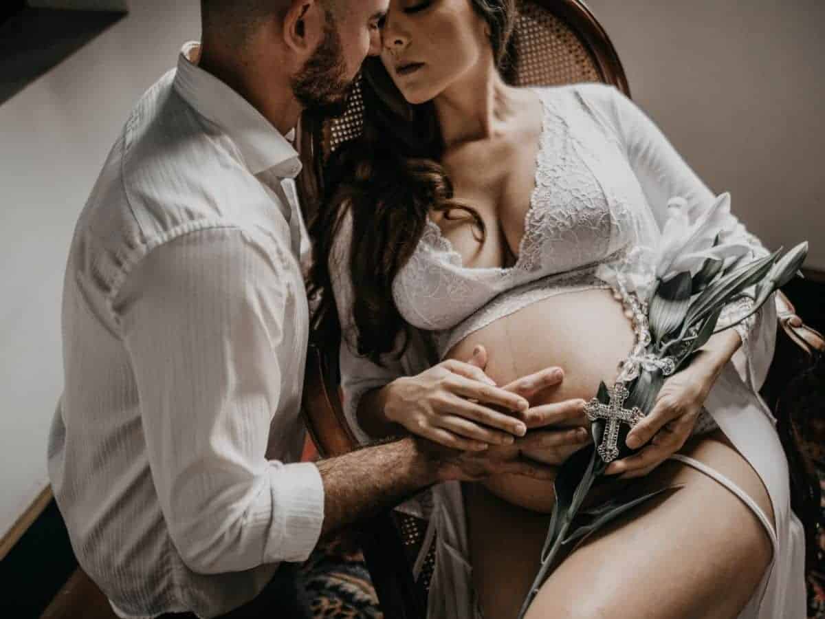 Pregnant woman sitting on a chair and holding flowers with her partner touching her belly.