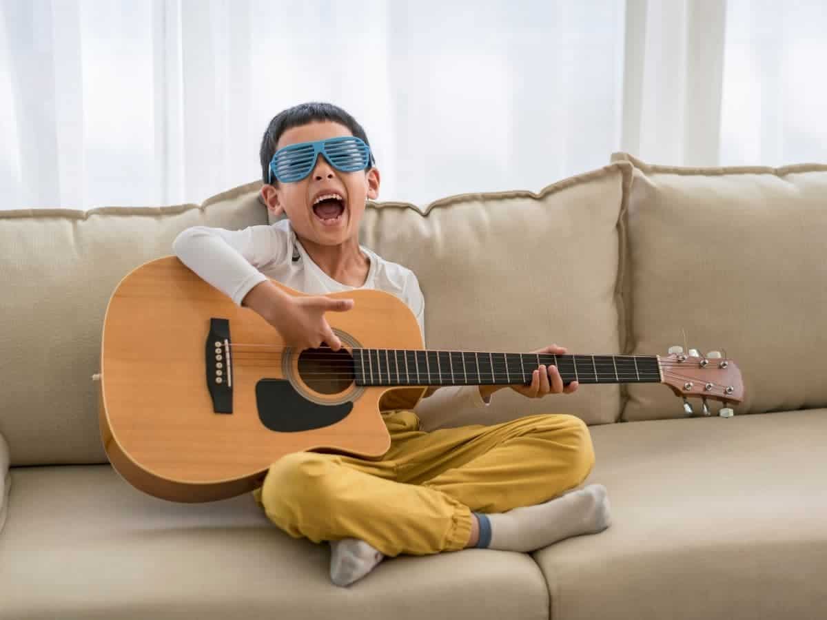 Kid sitting on a couch and playing a guitar while wearing sunglasses.