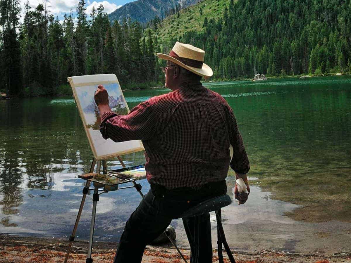 Person painting near a lake, trees, and mountains.
