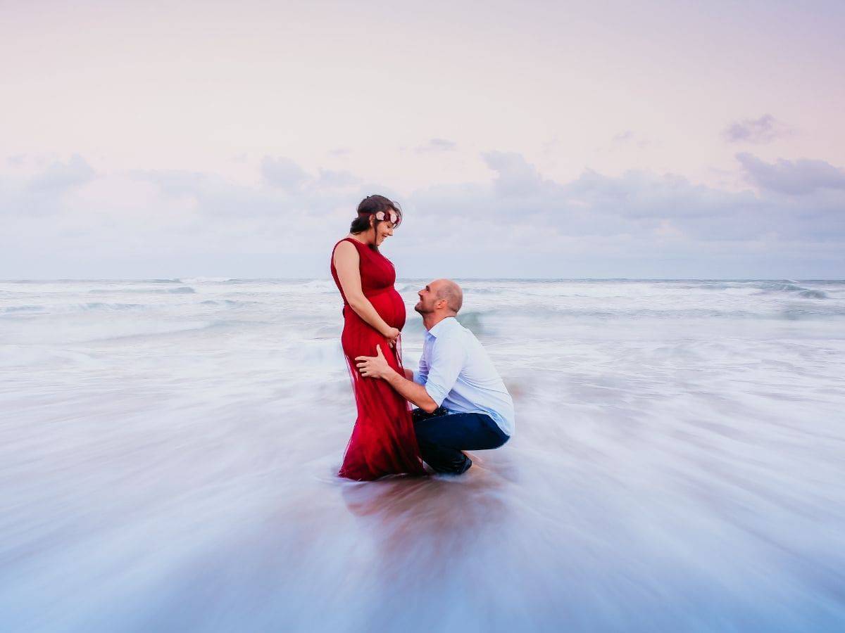 Pregnant woman standing and a man crouching down in front of her in the water.