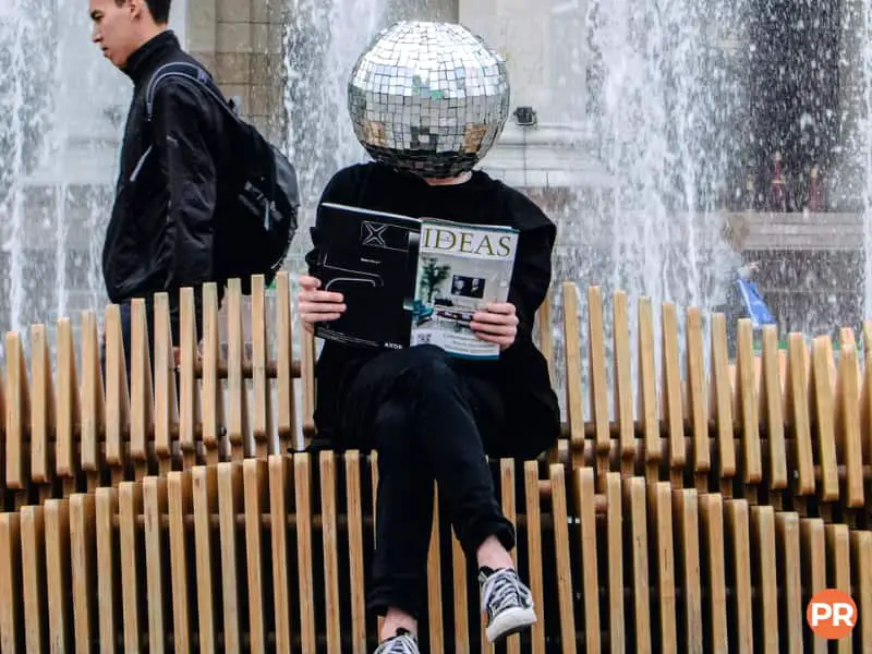 Person sitting on a bench wearing a reflective disco ball and reading a magazine.