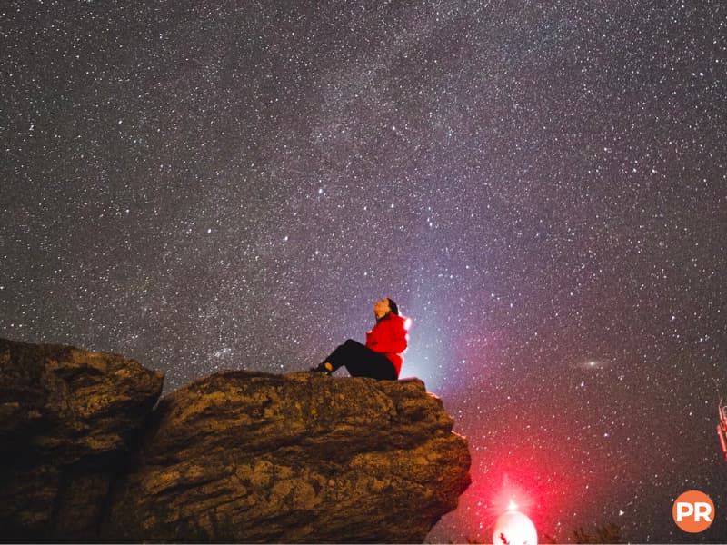 Person sitting on a cliff and looking up at the stars in the night sky.