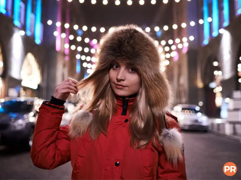 Person wearing a fur hat and standing under string lights at night.