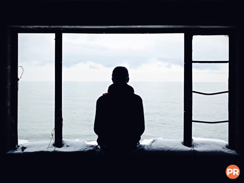 Silhouette of a person sitting on a ledge in front of the water.