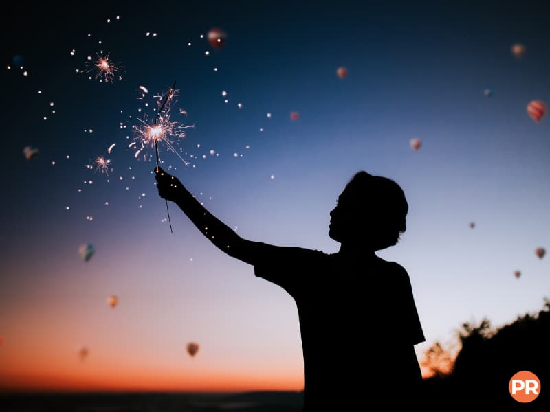 Silhouette of a person holding a sparkler with balloons in the sky.